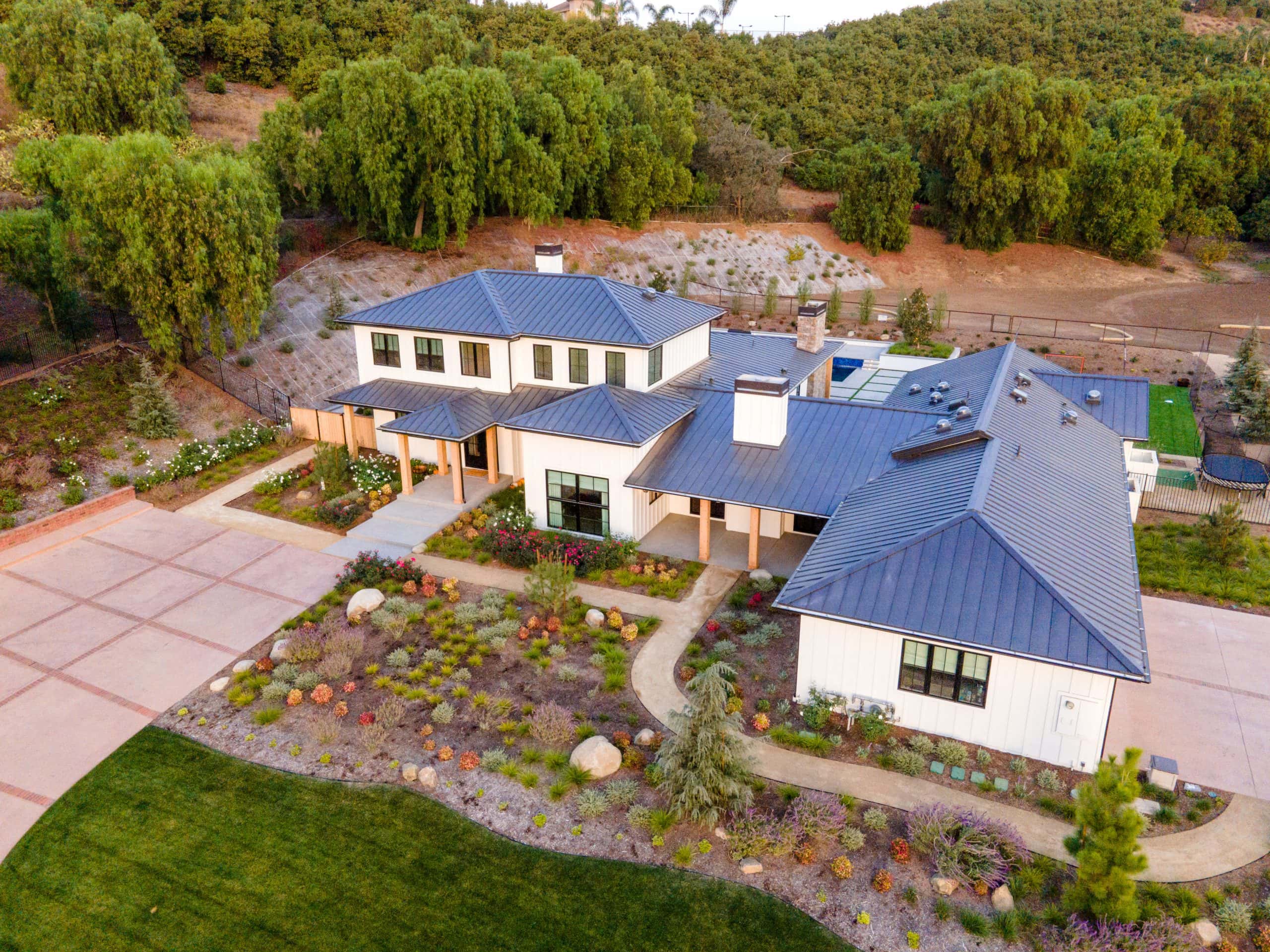 A modern farmhouse with blue metal roof, landscaped yard, and surrounding trees.