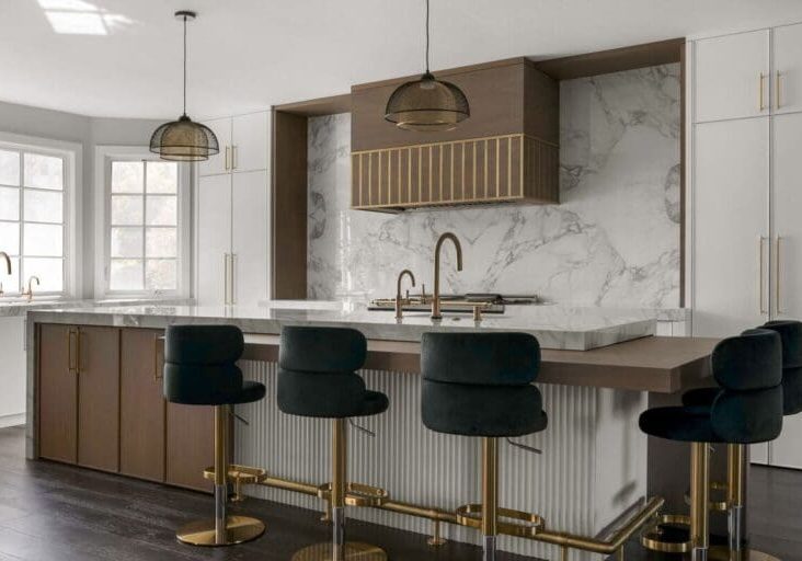 Modern kitchen with marble countertops, an island with dark green velvet bar stools, brass fixtures, white and wood cabinetry, pendant lights, and large windows providing natural light. This space exudes sophisticated glamour at every turn.